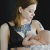 Calm female in black lingerie holding baby in arms and breastfeeding looking at infant against black background in photo studio