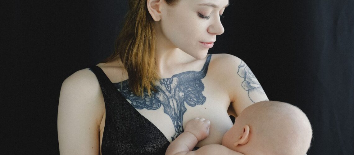 Calm female in black lingerie holding baby in arms and breastfeeding looking at infant against black background in photo studio