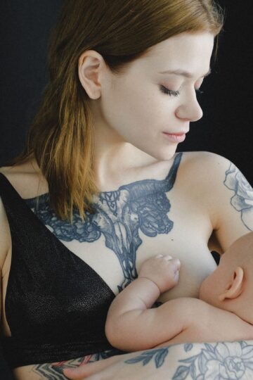 Calm female in black lingerie holding baby in arms and breastfeeding looking at infant against black background in photo studio