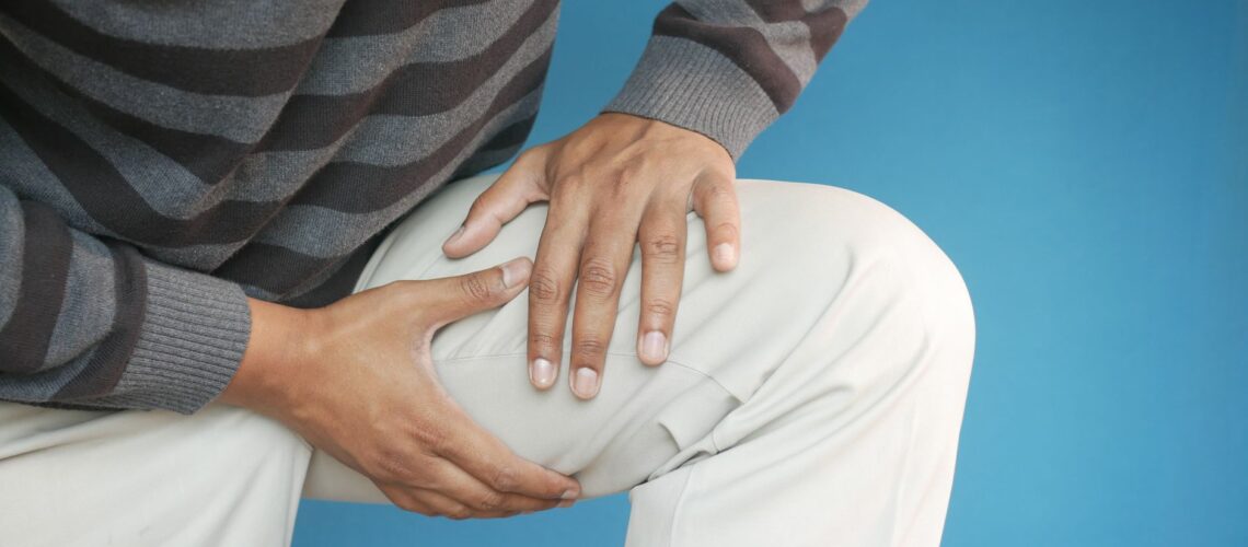 Close-up of a man's hands on his knee, showing pain or injury, on a blue background.
