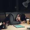 Man in office suit sleeping on desk with coffee and tablet nearby, symbolizing work fatigue.