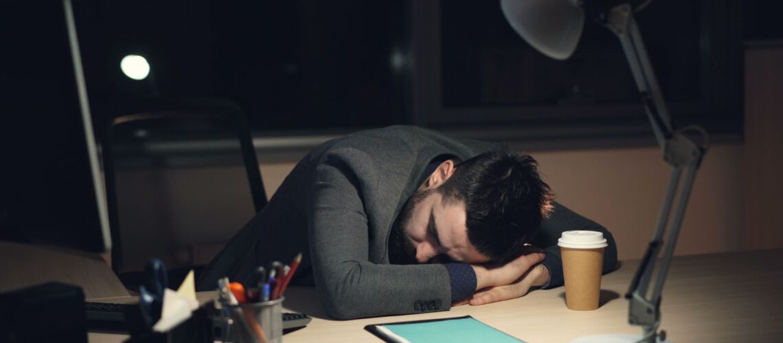Man in office suit sleeping on desk with coffee and tablet nearby, symbolizing work fatigue.