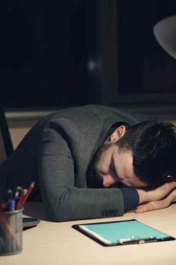 Man in office suit sleeping on desk with coffee and tablet nearby, symbolizing work fatigue.