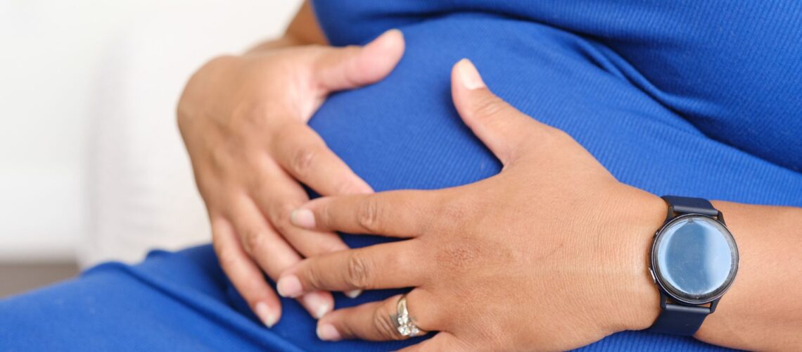 Closeup of a pregnant woman resting her hands on her belly, displaying a watch and a ring.