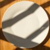 An empty white plate casts dramatic shadows on a bamboo mat in sunlight.
