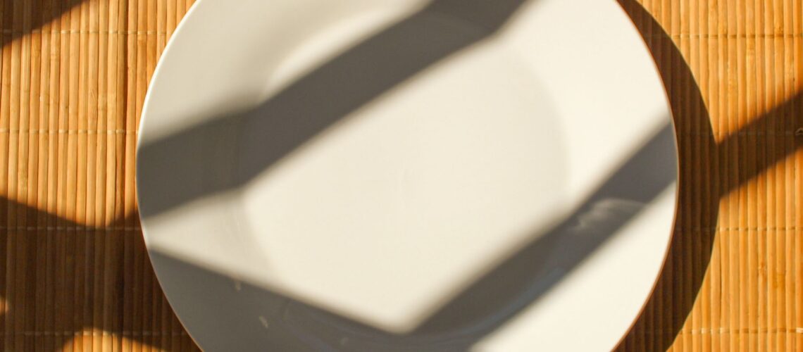 An empty white plate casts dramatic shadows on a bamboo mat in sunlight.