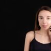Teenage girl with acne applying skincare cream on face, isolated black background.