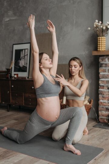 Pregnant woman doing yoga poses with a trainer in cozy indoor setting.