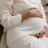 A serene moment of a pregnant woman in a cozy white dress holding her belly.