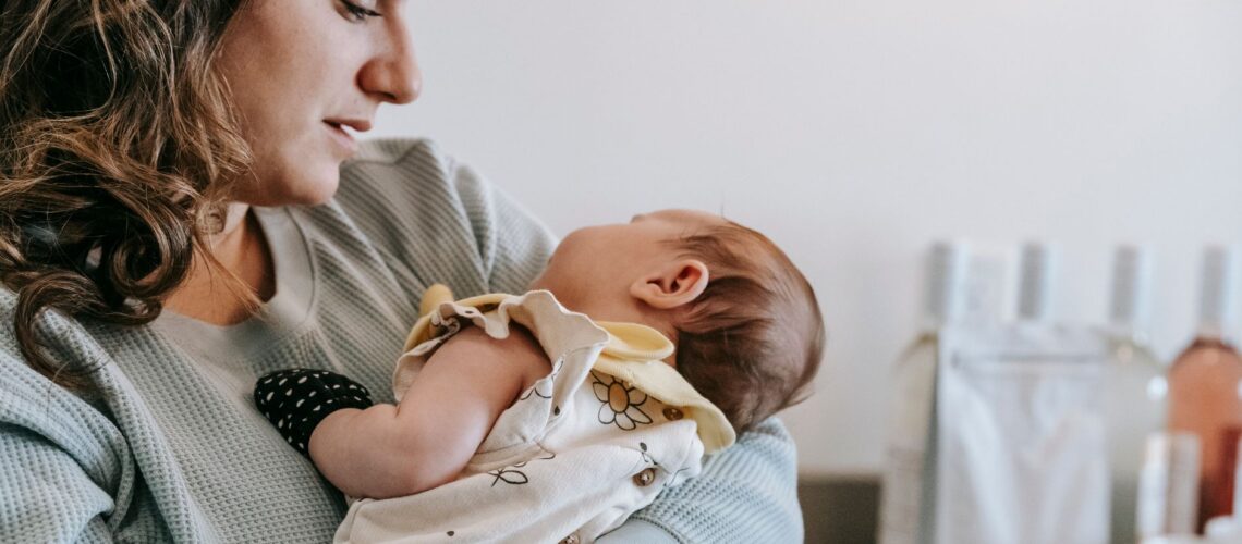 Side view of crop caring mother in casual wear with cute newborn baby in arms standing in light room at home