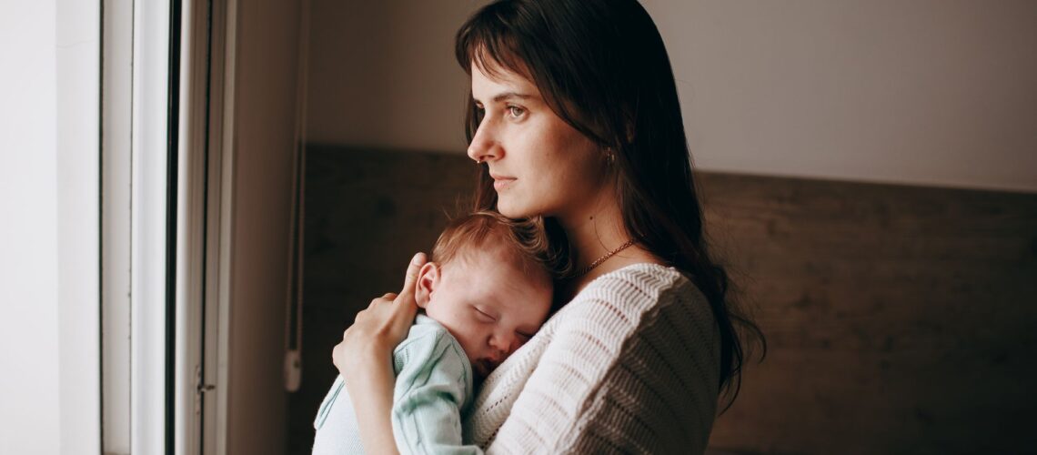 A serene moment of a mother holding her sleeping baby near a window, indoors.