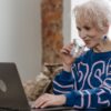 Elderly woman sipping water while working on a laptop in a cozy indoor setting.