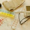 Flat lay image of healthcare essentials including a blood pressure monitor, pills, and eyeglasses on a white surface.
