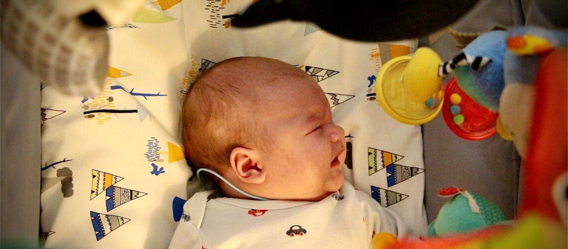 Crying newborn baby lying in a crib surrounded by colorful toys in a cozy setting.
