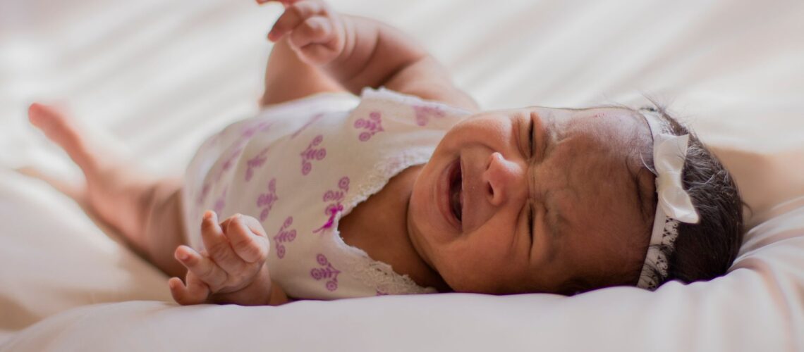 A close-up of a crying baby wearing a headband, lying on a soft bed indoors.