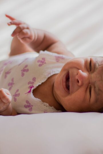 A close-up of a crying baby wearing a headband, lying on a soft bed indoors.