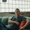 A person with green hair relaxing on a teal sofa in a chic industrial indoor setting.