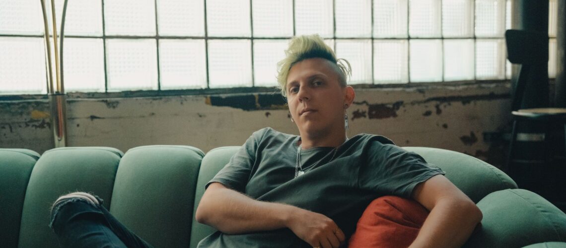 A person with green hair relaxing on a teal sofa in a chic industrial indoor setting.