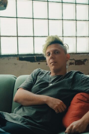 A person with green hair relaxing on a teal sofa in a chic industrial indoor setting.