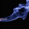 Detailed close-up of a cigarette with glowing ember and swirling smoke on a black background.