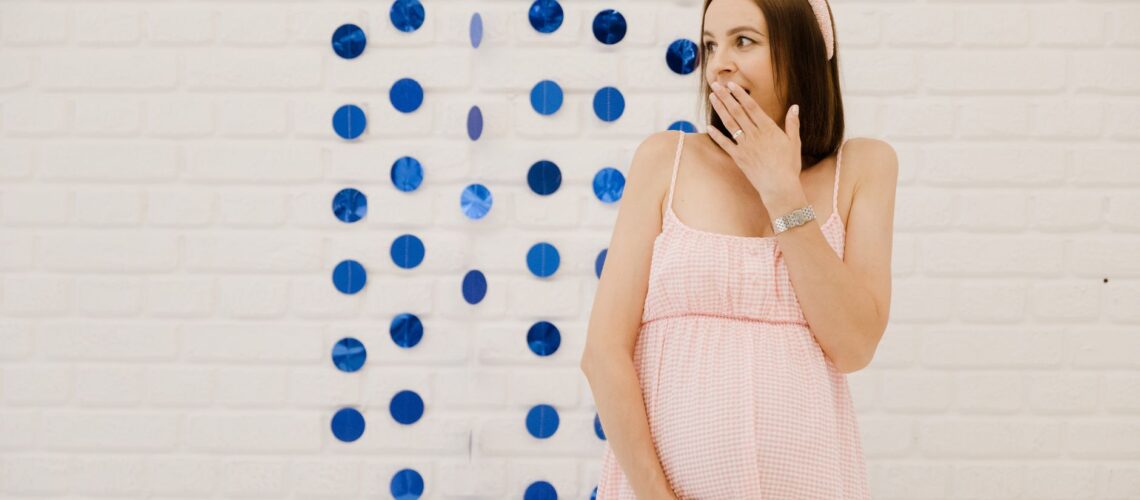 A surprised pregnant woman in a pink dress at a gender reveal party indoors with blue decorations.