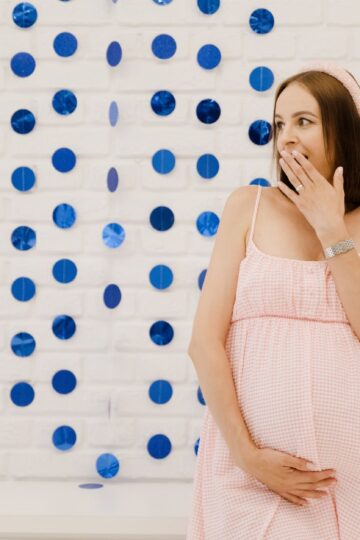 A surprised pregnant woman in a pink dress at a gender reveal party indoors with blue decorations.