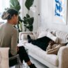 Ethnic female therapist with clipboard sitting near male client lying on couch with clasped hands during psychotherapy session in cozy office
