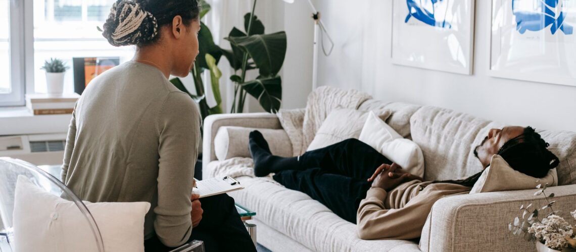 Ethnic female therapist with clipboard sitting near male client lying on couch with clasped hands during psychotherapy session in cozy office