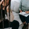 Crop concerned African American male sitting on sofa with female psychologist and holding head in hands in dismay during psychotherapy session