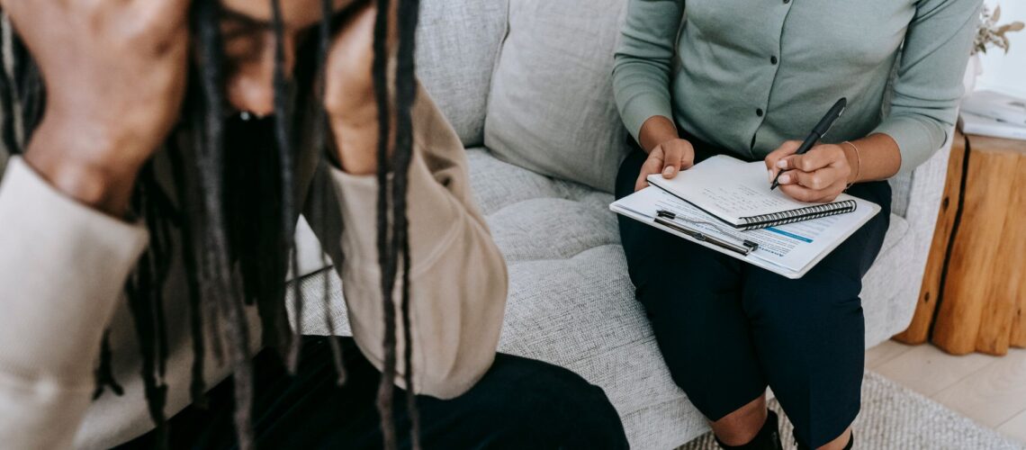 Crop concerned African American male sitting on sofa with female psychologist and holding head in hands in dismay during psychotherapy session
