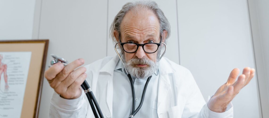 Senior doctor gesturing with stethoscope in a clinical setting, emphasizing telemedicine.