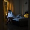 Woman wrapped in blanket watching TV, battling insomnia in dimly lit bedroom.