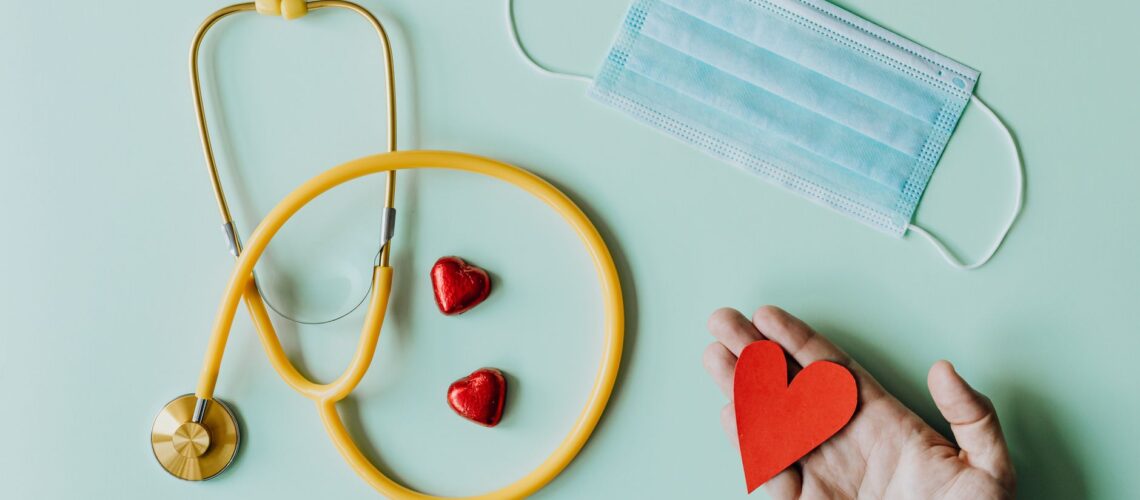 Top view of crop anonymous person hand with red paper heart on table with stethoscope and medical mask for coronavirus prevention