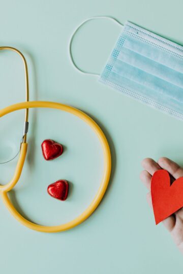 Top view of crop anonymous person hand with red paper heart on table with stethoscope and medical mask for coronavirus prevention