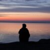 A contemplative silhouette of a person sitting by the calm sea during a beautiful sunset.