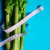 Close-up of celery stalks wrapped with measuring tape on a vibrant blue background, symbolizing health and diet.