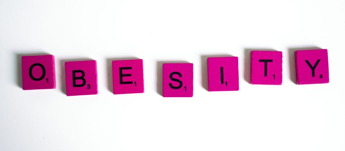 Purple Scrabble tiles spelling 'obesity' on a white surface, conceptual still life.