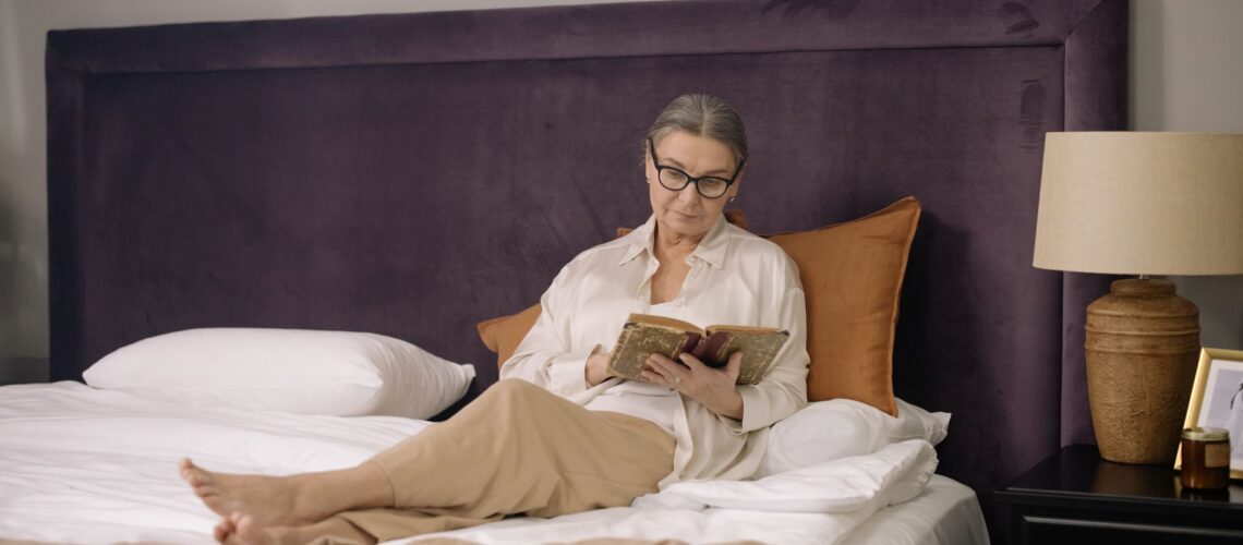 A senior woman enjoys reading a book in a cozy and elegant bedroom setting.