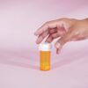A hand reaches for a prescription bottle with yellow pills on a pink background, symbolizing healthcare.