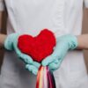 A nurse in gloves holds a red heart with rainbow strings, symbolizing love and healthcare.