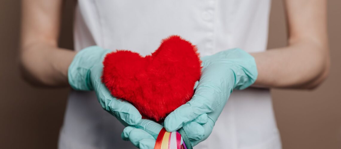 A nurse in gloves holds a red heart with rainbow strings, symbolizing love and healthcare.