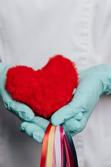 A nurse in gloves holds a red heart with rainbow strings, symbolizing love and healthcare.