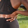 Woman in athletic attire holding her back outside, indicating discomfort or pain.