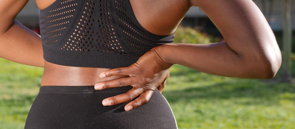 Woman in athletic attire holding her back outside, indicating discomfort or pain.