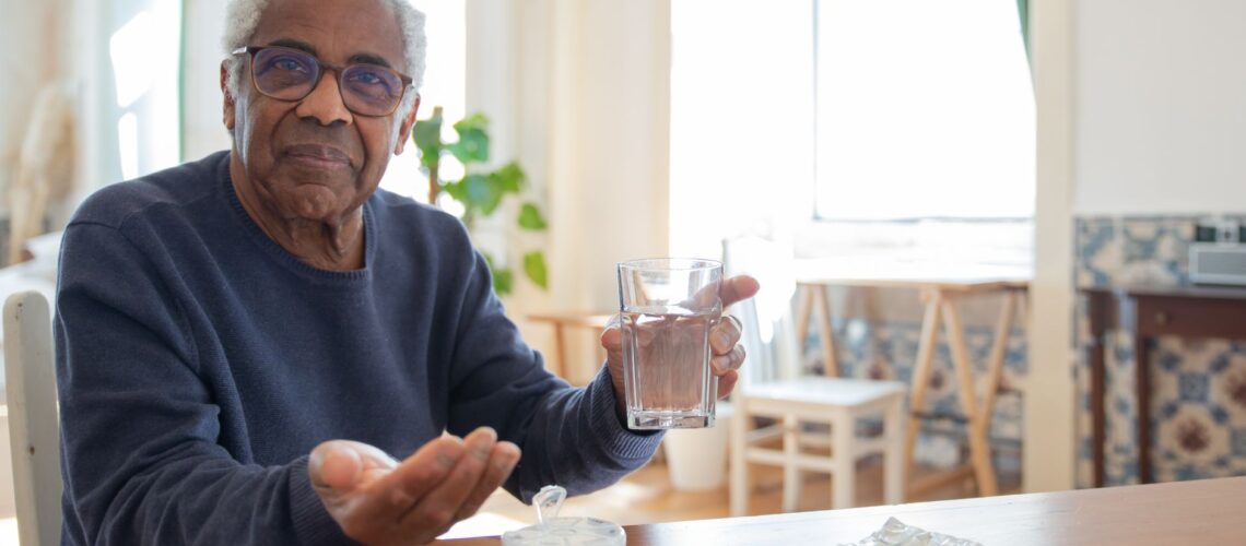 Elderly man sitting at a table at home, holding a glass of water and medication, emphasizing healthcare routine.