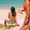 Two women in bikinis relaxing on a tropical beach, applying sunscreen.