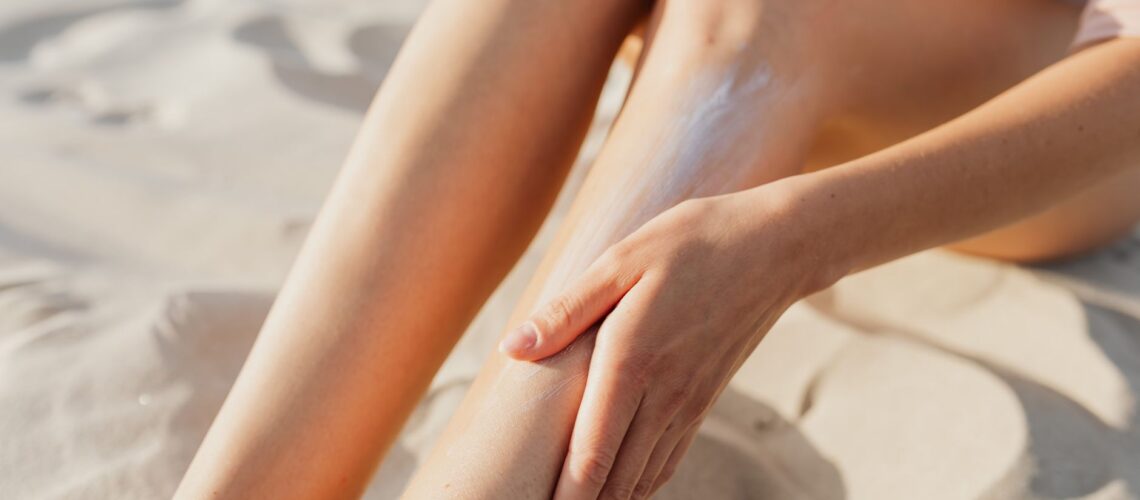 Close-up of a woman applying sunscreen on her legs while sitting on the beach sand.