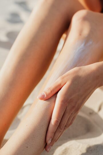 Close-up of a woman applying sunscreen on her legs while sitting on the beach sand.