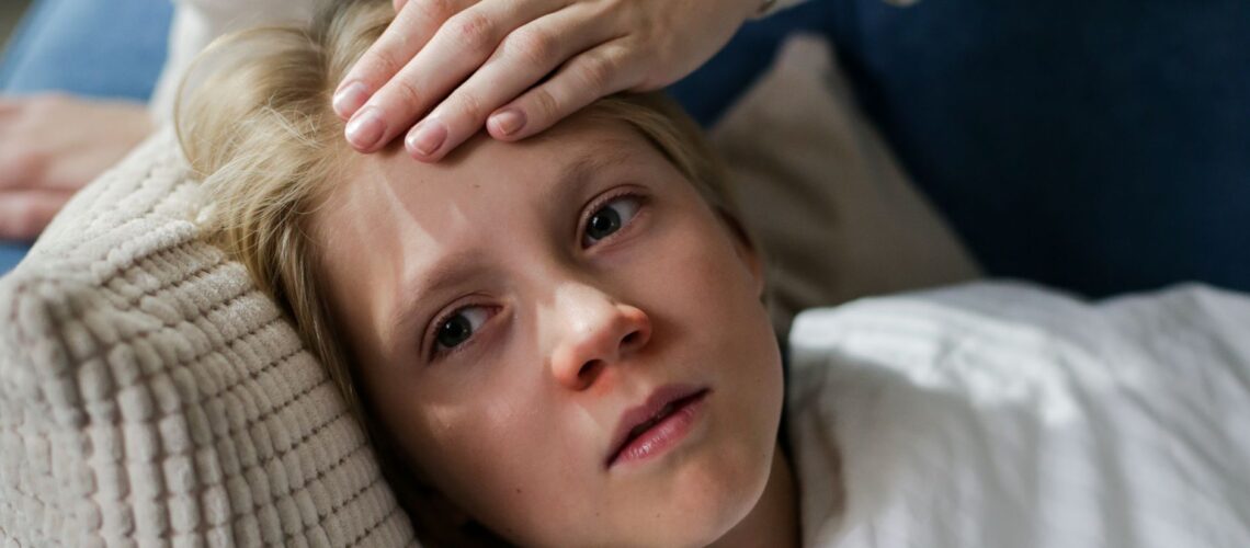 Young child lying down with a hand checking for fever, indicating illness or flu symptoms.