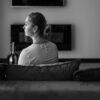 A senior woman sitting pensively on a couch, back view in a grayscale setting.